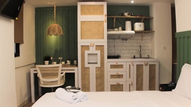 A compact hotel room with a bed, a desk, a chair, a kitchenette, and a hanging light fixture. - Minimalist Design Studio At Meikarta Apartment (Cikarang)