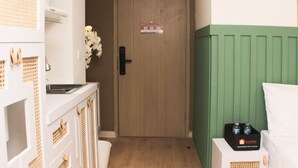 A hotel room with a wooden door, a green wall, a white bed, and a small table with a bottle of water. - Minimalist Design Studio At Meikarta Apartment (Cikarang)