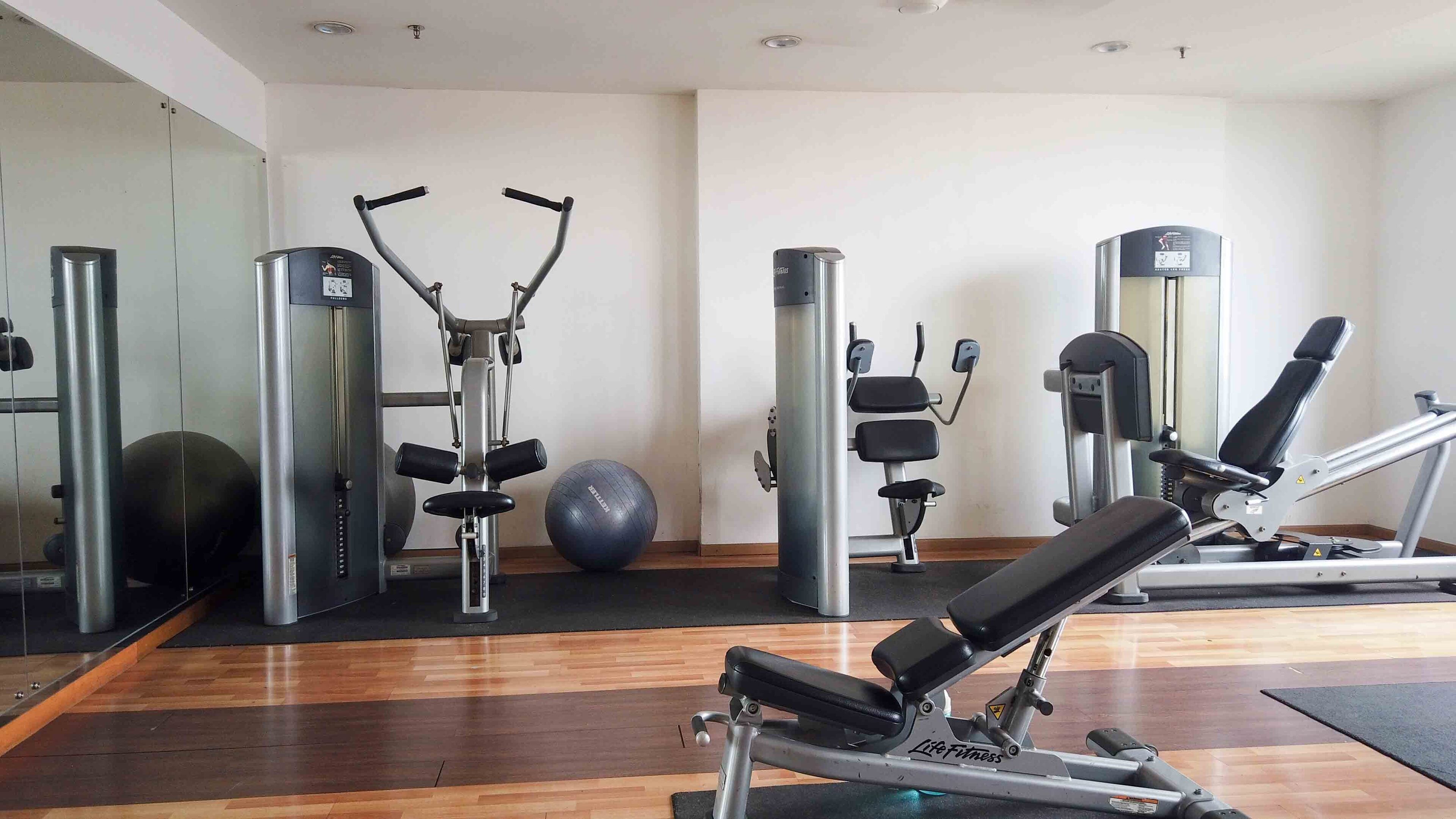 Academia com equipamentos de ginástica, espelho e piso de madeira.