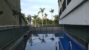 A swimming pool with metal railings and a blue tiled floor, surrounded by buildings and palm trees. - Homey Furnished Studio At Beverly Dago Apartment (Bandung)