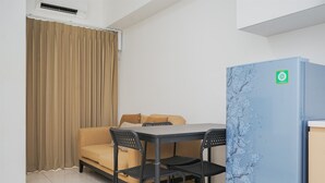 Ein Zimmer mit einem beigen Sofa, einem kleinen Esstisch mit Stühlen, einem blauen Kühlschrank und einem Fenster mit beigen Vorhängen.