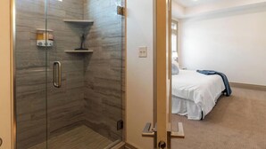 A modern bathroom with a glass-enclosed shower, a ceiling fan, and a visible bed in the adjacent room. - BRAND NEW The Townie (Cedar City)