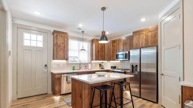 A modern kitchen with wooden cabinets, stainless steel appliances, and a central island with stools. - BRAND NEW The Townie (Cedar City)