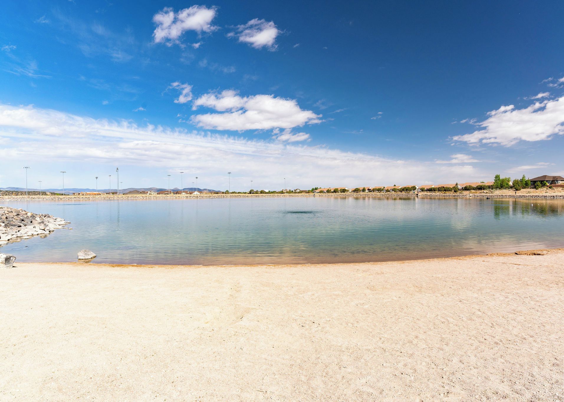 A calm lake with a sandy shore, surrounded by a clear blue sky and distant hills.
