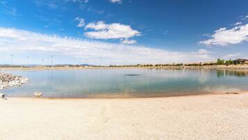 A calm lake with a sandy shore, surrounded by a clear blue sky and distant hills.