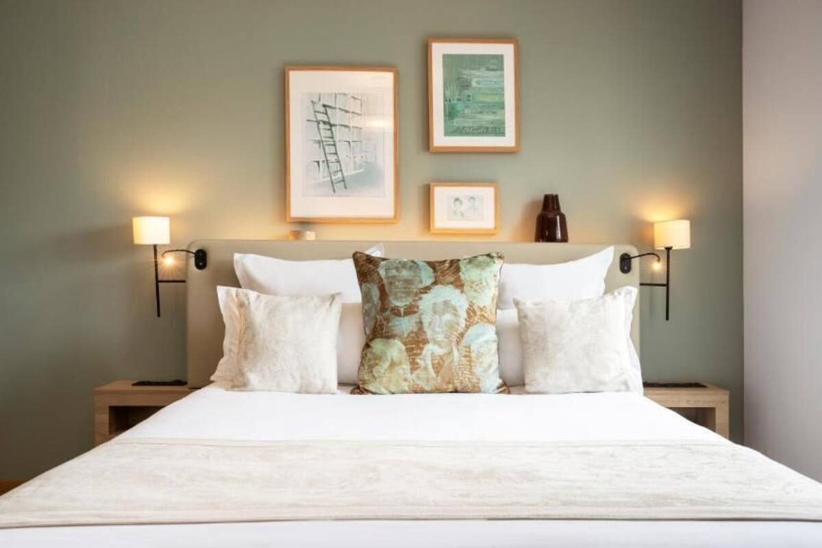 Una cama con ropa de cama blanca, dos mesitas de noche, lámparas fijadas a la pared y una obra de arte enmarcada.