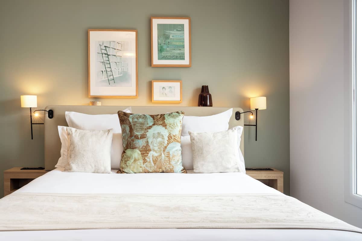 Una cama bien hecha con sábanas blancas y un cojín decorativo, flanqueada por dos mesitas de noche con lámparas de pared.