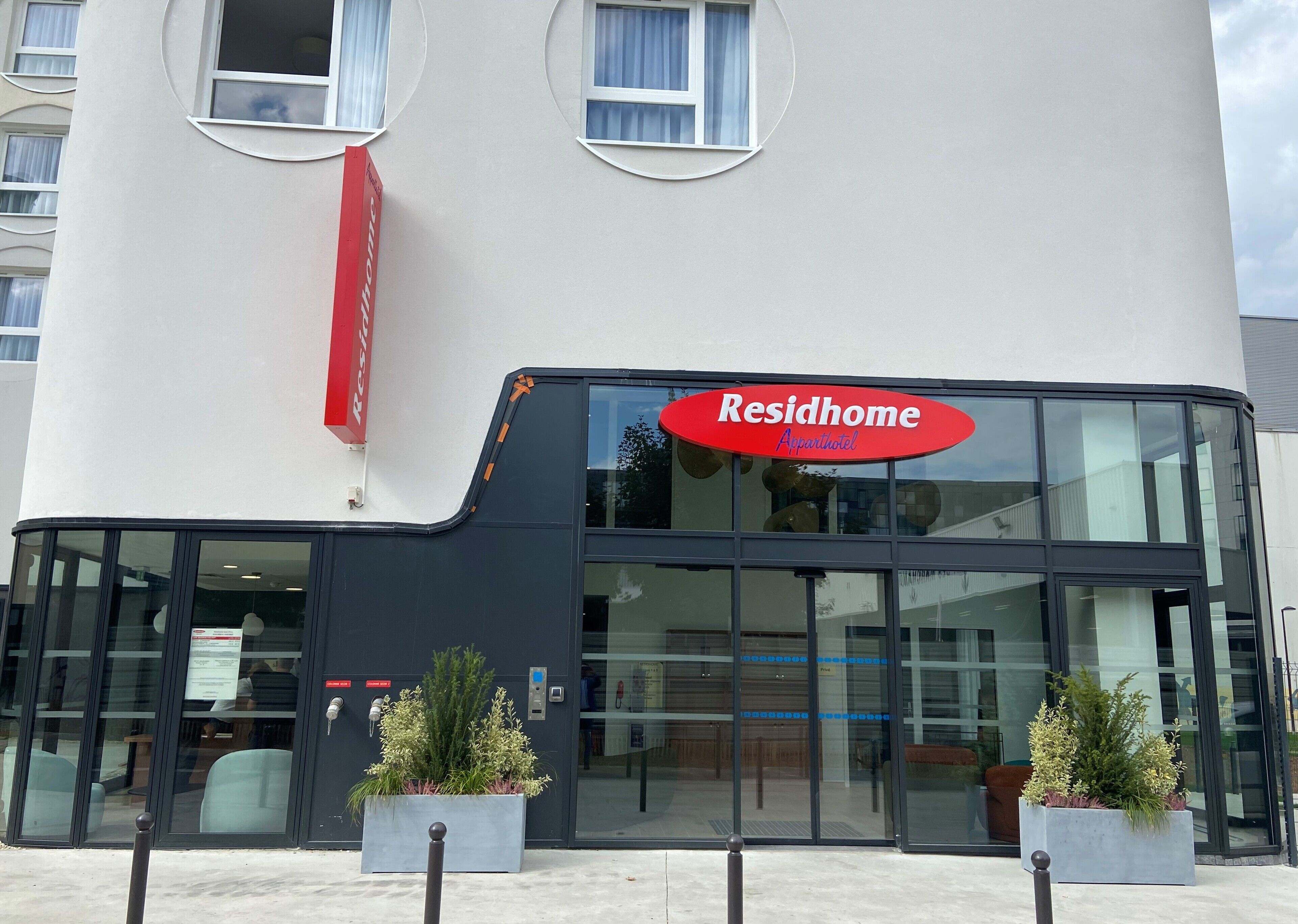 A building with a large glass facade, a red sign reading "Residhome Apartmenthotel," and two potted plants in front.