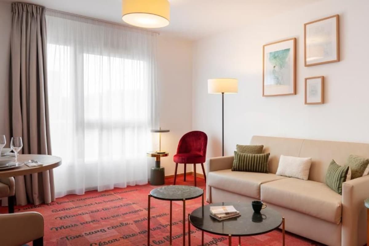 A hotel room with a sofa, a red chair, a small table, a floor lamp, and a dining table with glasses.