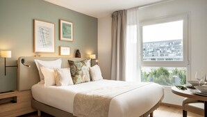 A hotel room with a large bed, bedside lamps, framed pictures on the wall, a window with a view of buildings, and a small table with glasses and a bowl. - Residhome Quai d'Ivry (Ivry-sur-Seine)