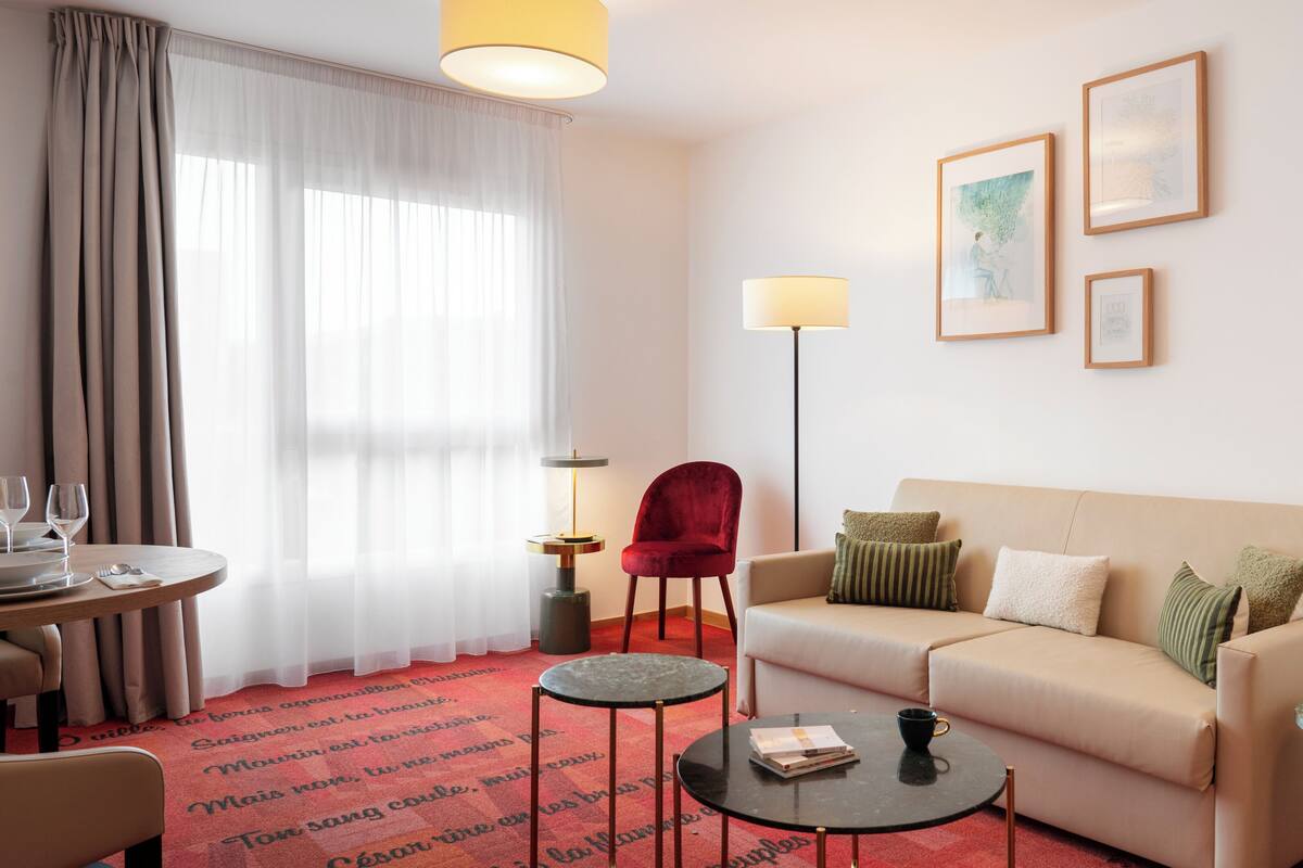 Una habitación de hotel moderna con un sofá color beige, una silla roja, una mesa redonda pequeña y una zona de comedor con una mesa preparada para dos.