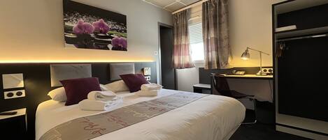 Habitación de hotel con una cama bien hecha, un escritorio con lámpara y un cuadro de flores en la pared.