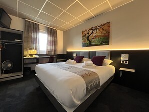 Une chambre d’hôtel avec un grand lit, un bureau, une chaise, un ventilateur et un tableau au mur.