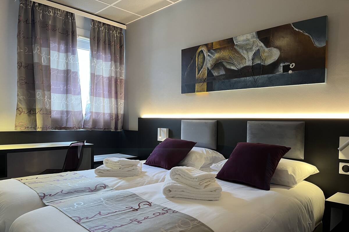 Ein Hotelzimmer mit einem Bett, zwei Kissen, einem Schreibtisch, einem Stuhl, einem Bild an der Wand und einem Fenster mit Vorhängen.