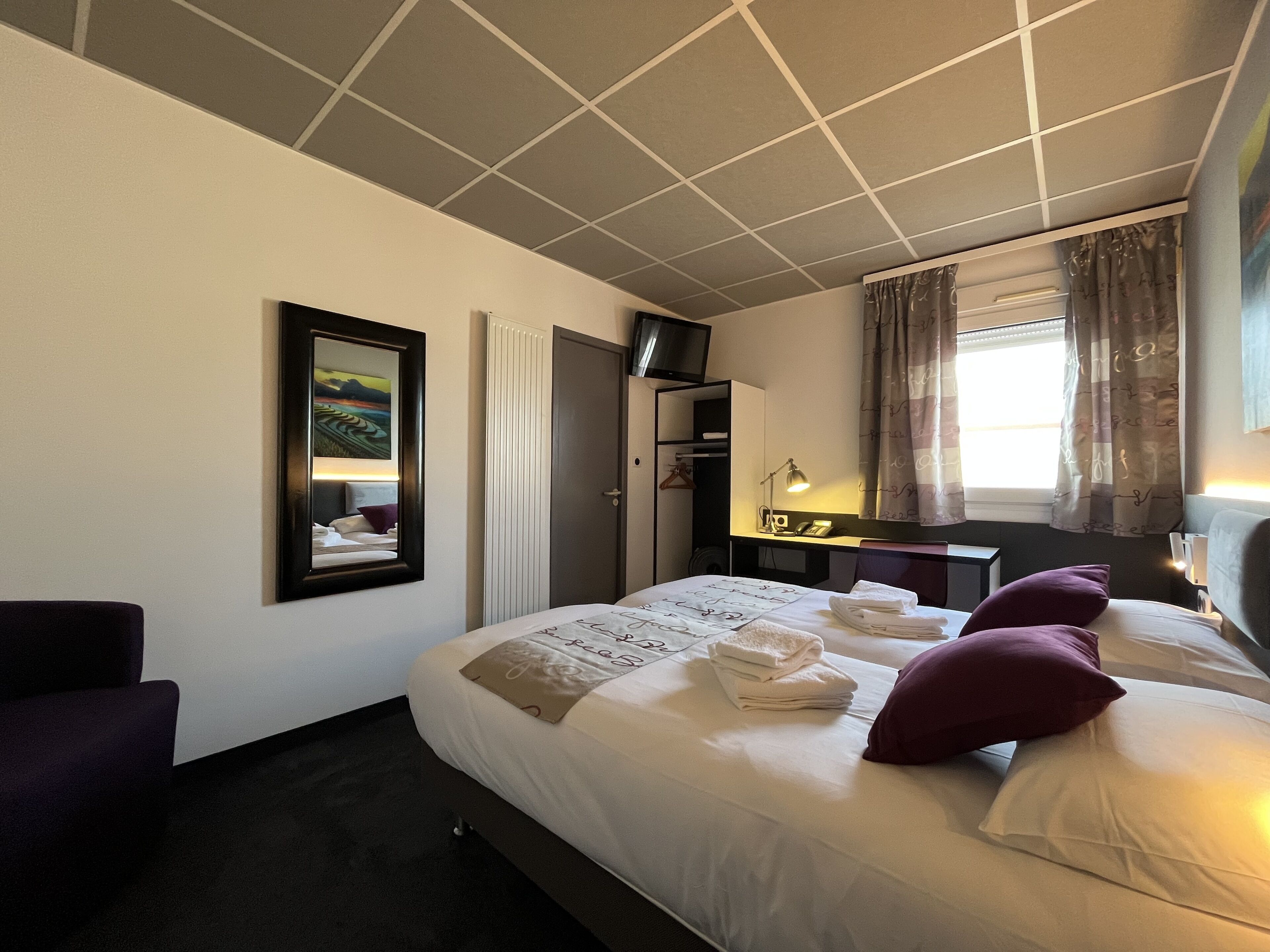A hotel room with a bed, a desk, a chair, a TV, a wardrobe, and a window with curtains.