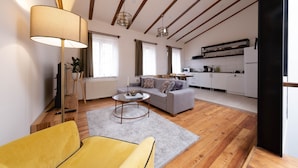 Un salon moderne avec un canapé, un fauteuil jaune, une table basse et une lampe. Au fond, on aperçoit une cuisine avec des meubles blancs et un plan de travail noir.