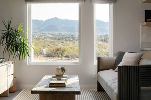 A modern living room with a large window, a sofa, a coffee table with books, and a plant.