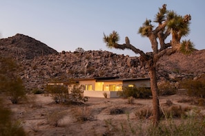 A desert house with a rocky hill in the background, surrounded by sparse vegetation.