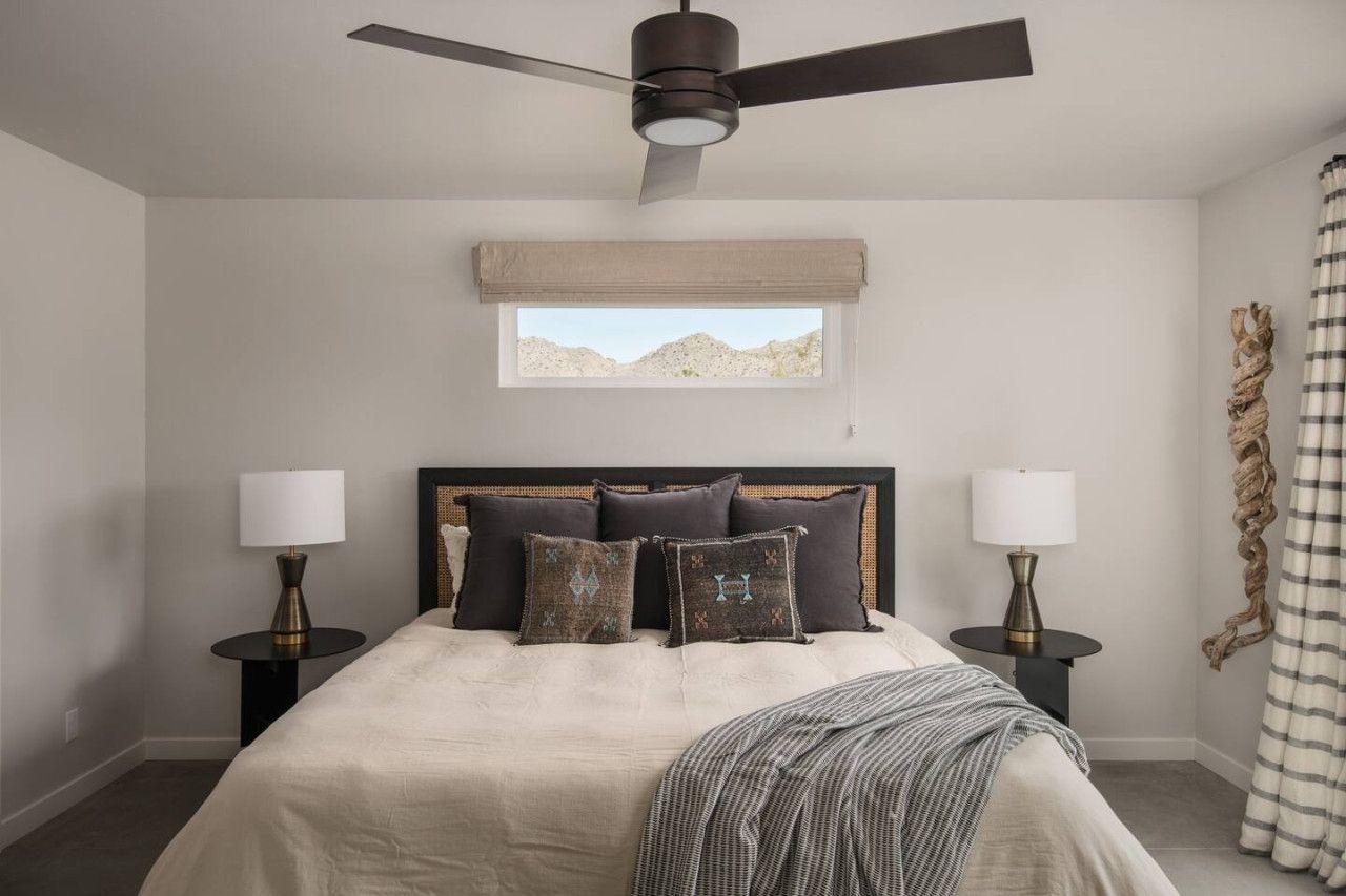 Quarto com cama, duas mesas de cabeceira, ventilador de teto, janela com vista e cortina.