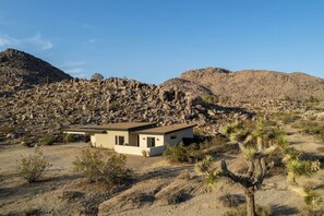 A modern house with a flat roof, surrounded by desert vegetation and rocky hills.