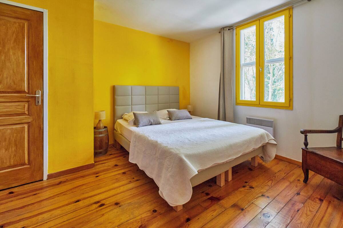 A bedroom with a wooden floor, a bed with a white bedspread, a wooden chair, and a window with yellow frames.