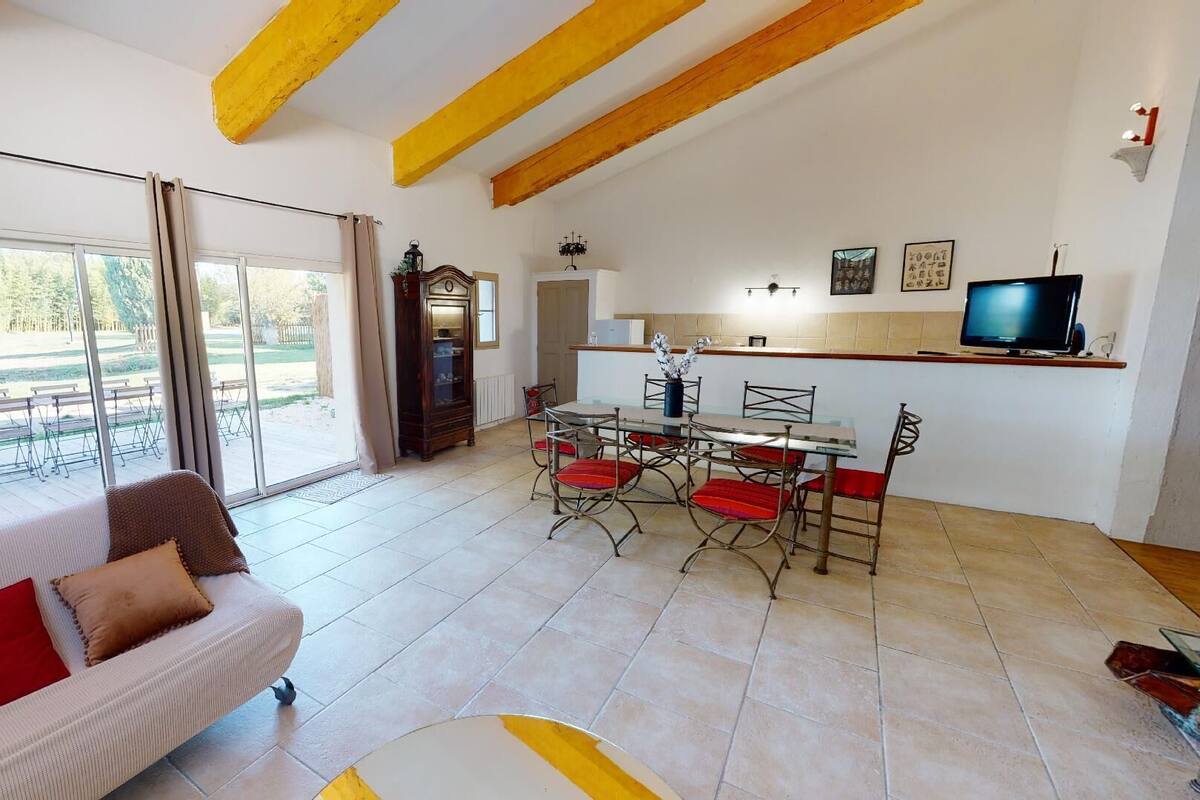 A spacious living area with a sofa, dining table, and chairs, a kitchen with a TV, and large windows offering a view of the outdoors.
