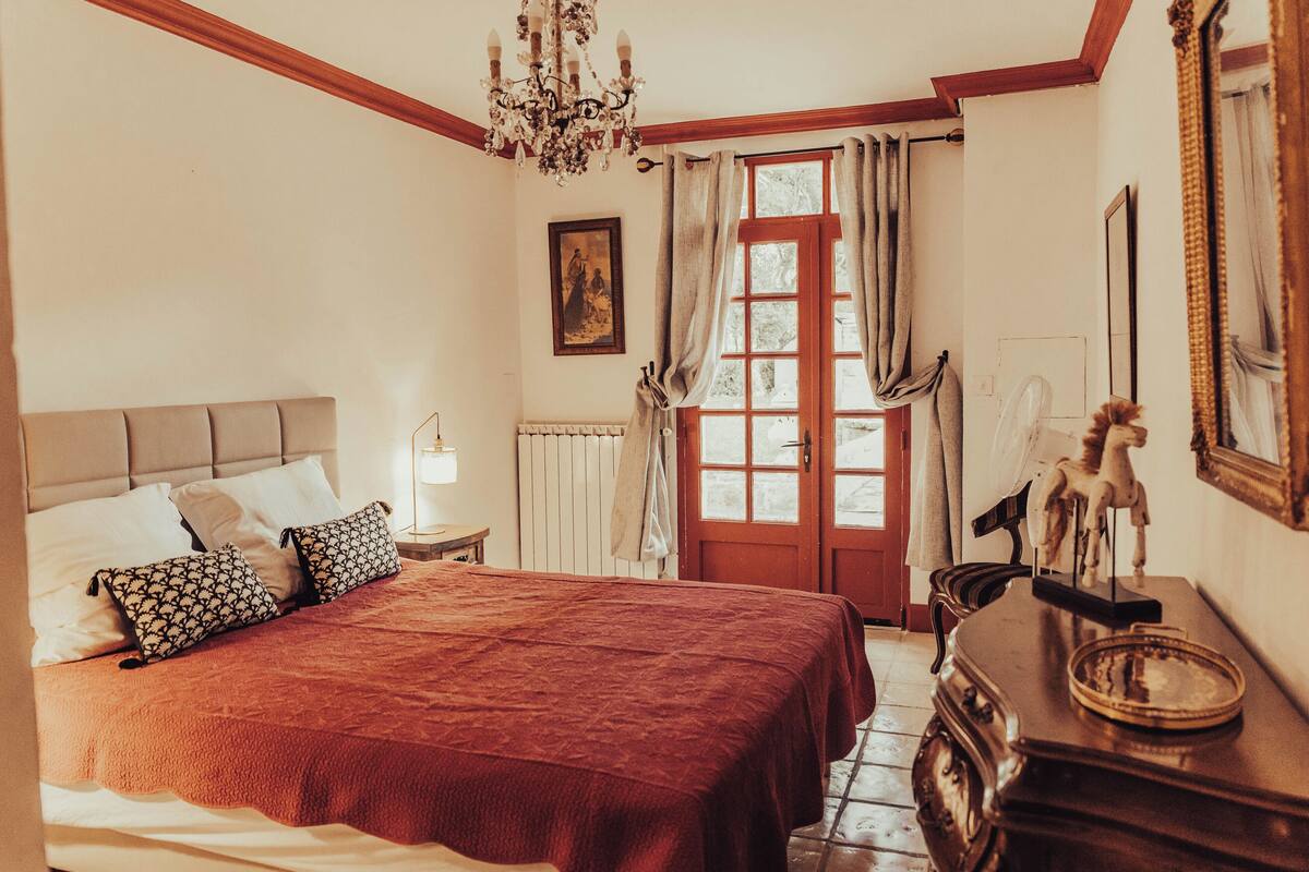 A bedroom with a large bed, a dresser, a chandelier, a door with curtains, and a framed picture on the wall.