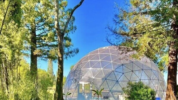 A geodesic dome structure with a small entrance, surrounded by a grassy area with trees and a pair of lounge chairs.