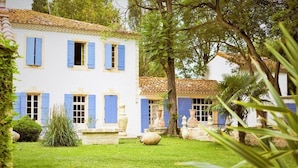 A white building with blue shutters and a tiled roof, surrounded by a well-maintained garden with grass, trees, and decorative pots.