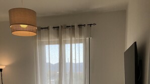 Ett sovrum med en säng, en stol, en tv och en balkong med tunna, genomskinliga gardiner.