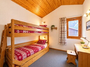 A bunk bed with red patterned bedding, a desk with a lamp, a window with curtains, and a small plant on the wall.