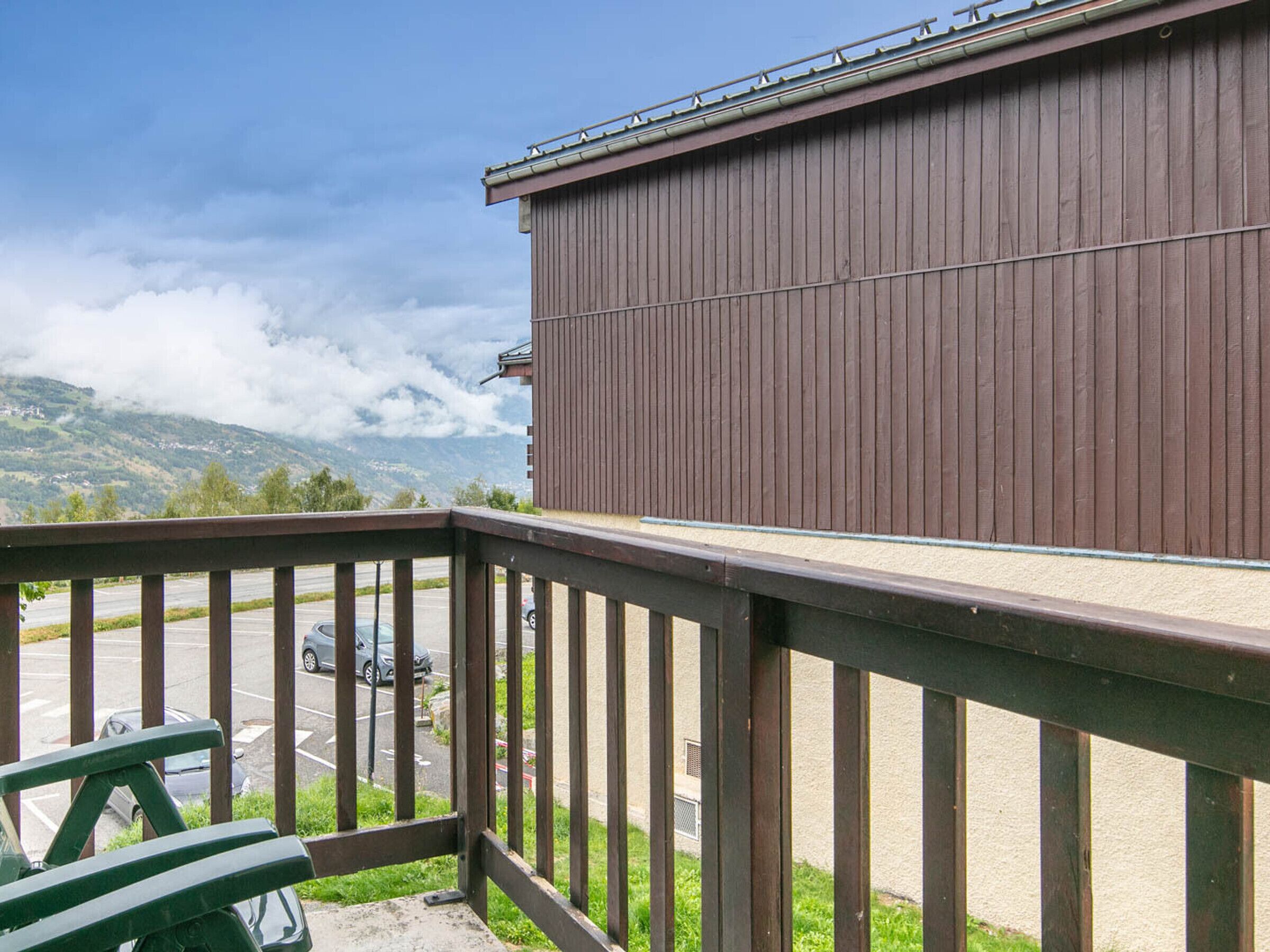 A view from a balcony with a wooden railing overlooking a parking area and a building with vertical siding.