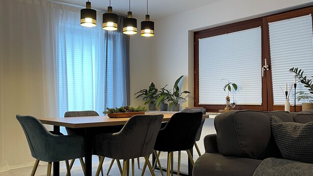 A modern dining area with a wooden table and chairs, a sofa, and a large window with blinds.
