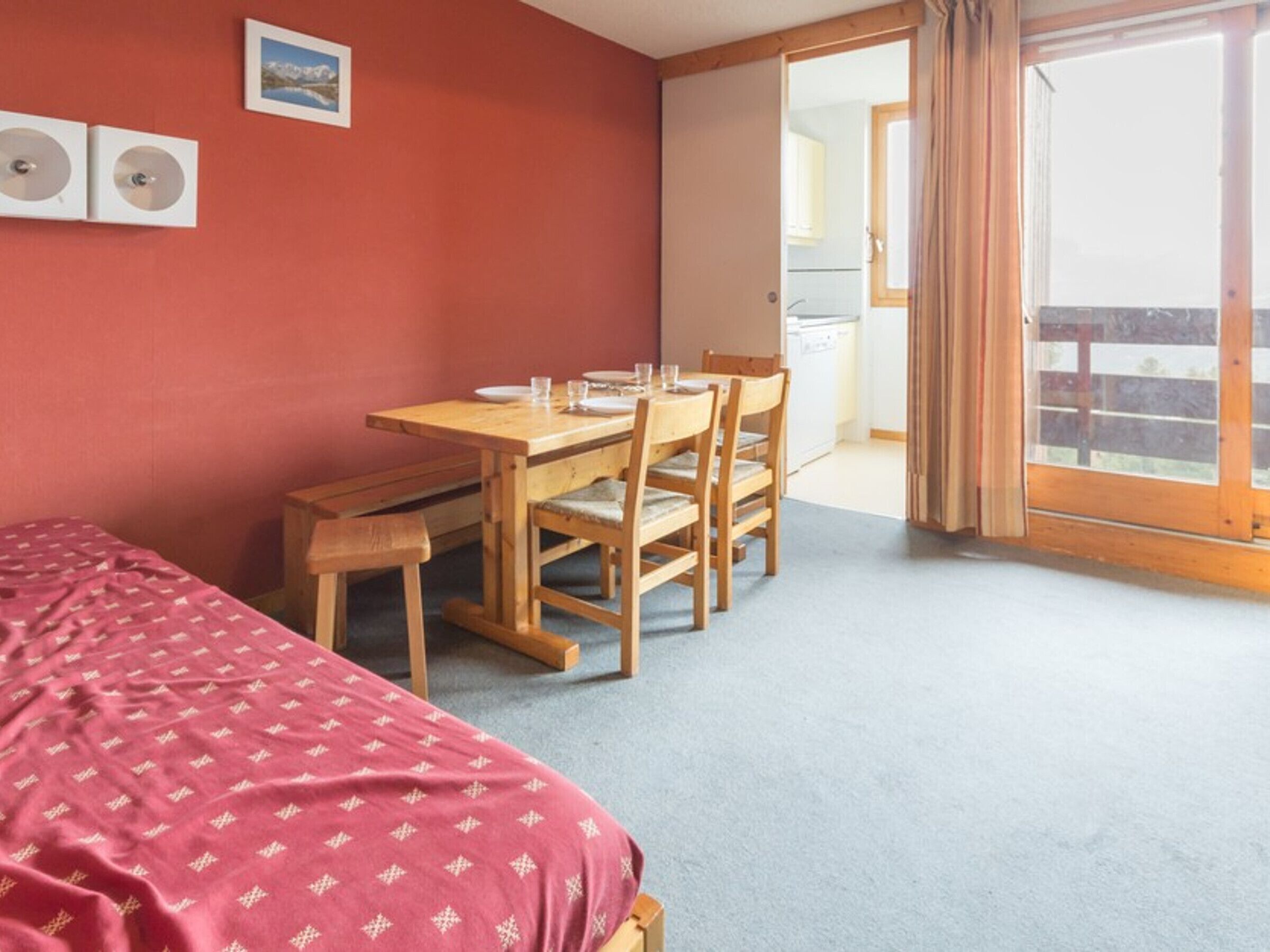 Quarto com uma cama, uma mesa de madeira com cadeiras, uma parede vermelha e vista para uma varanda.