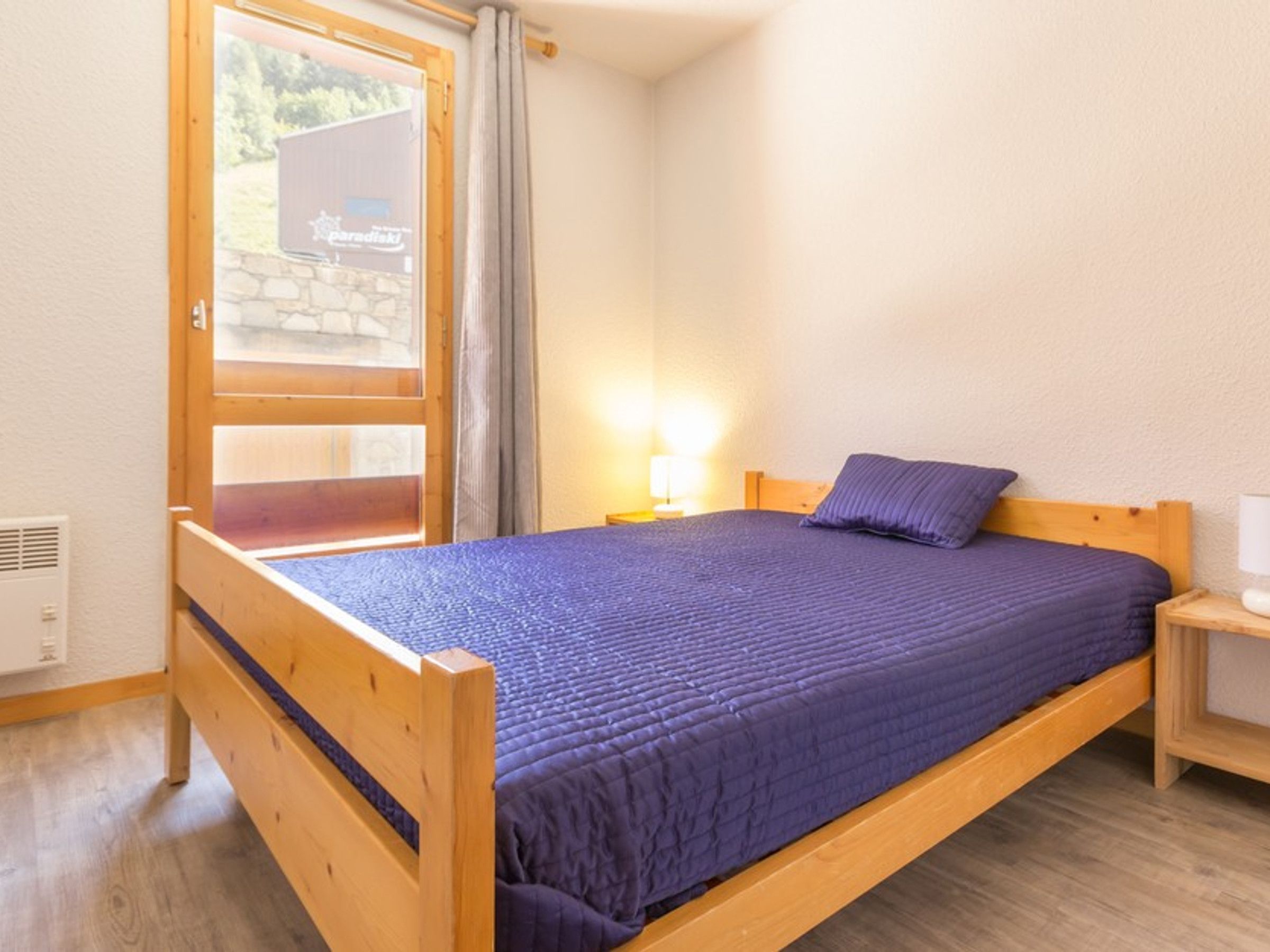 Habitación individual con cama de madera, colcha azul, mesita de noche con lámpara y ventana con vista a un edificio.