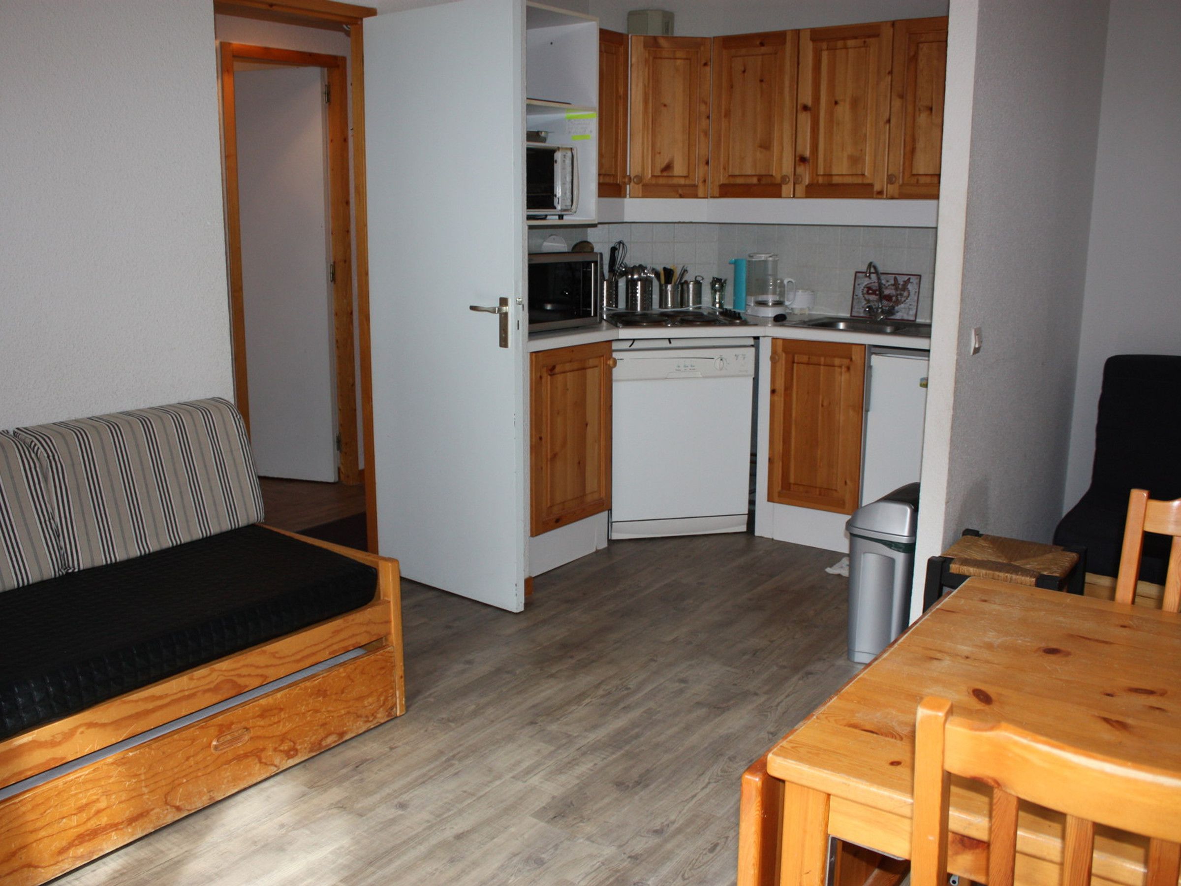 A compact kitchen with wooden cabinets, a dishwasher, and a microwave. There's a small dining table and a sofa with a black cushion.