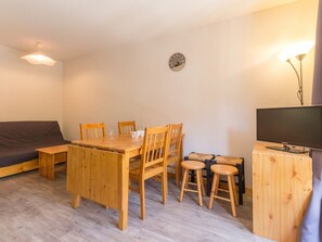 Un espacio habitable compacto con una mesa de madera, cuatro sillas, un sofá, un televisor sobre un soporte de madera y un reloj de pared.