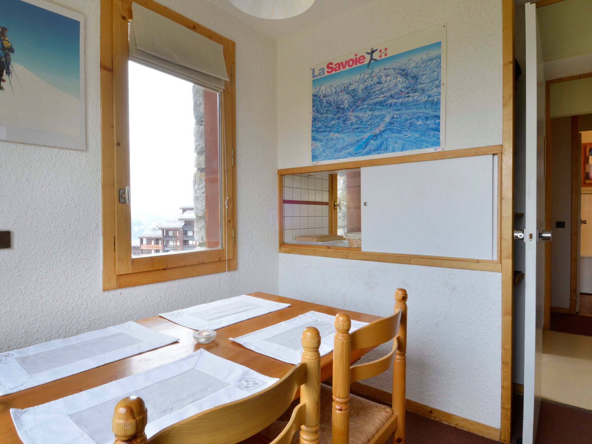 Een kamer met een houten tafel gedekt voor twee personen, een houten stoel, een raam met uitzicht op gebouwen en een poster aan de muur.