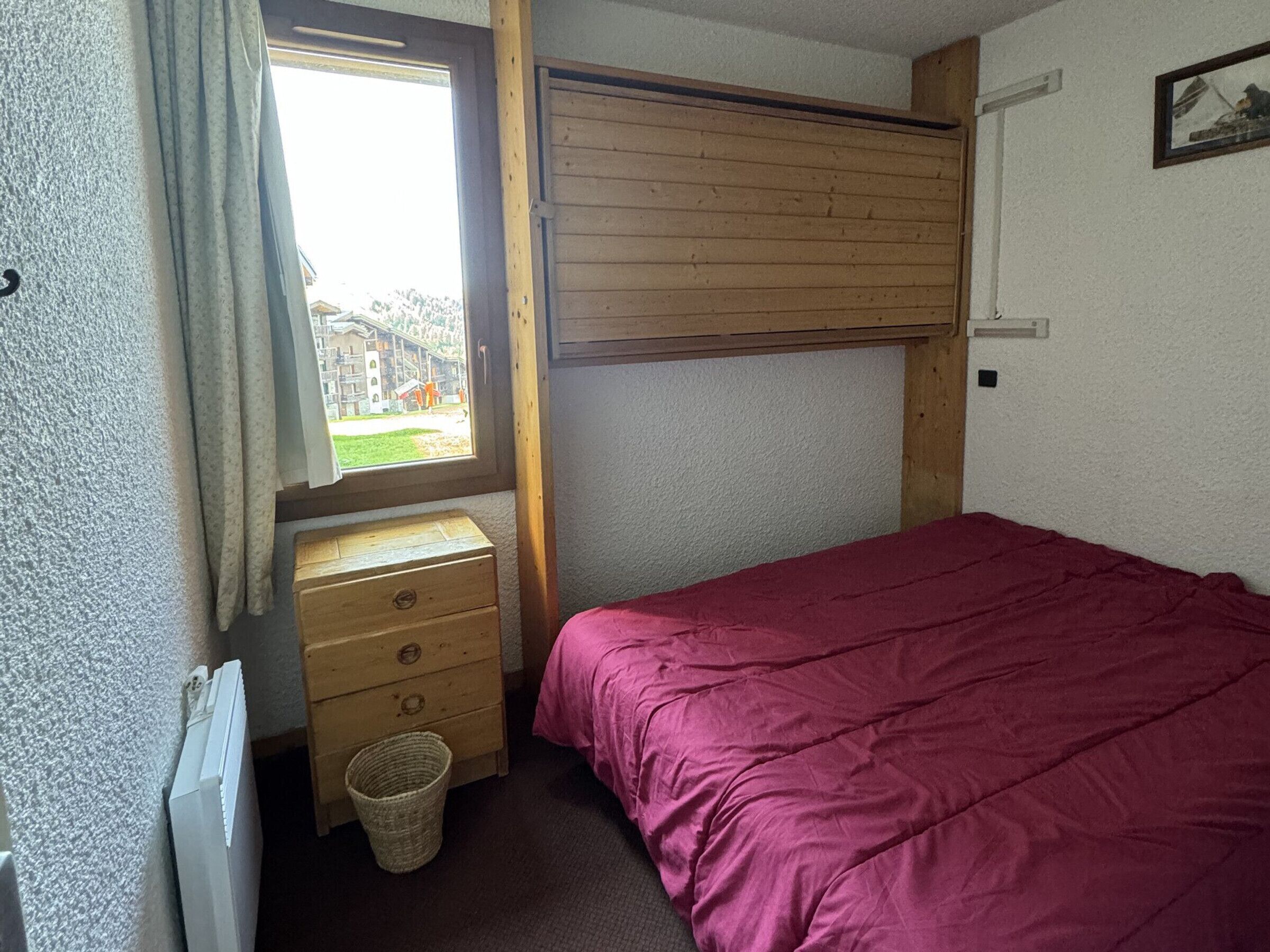 Ein kleines Schlafzimmer mit einem Einzelbett, einem Nachttisch aus Holz, einem Fenster mit Vorhängen und einer Heizung.