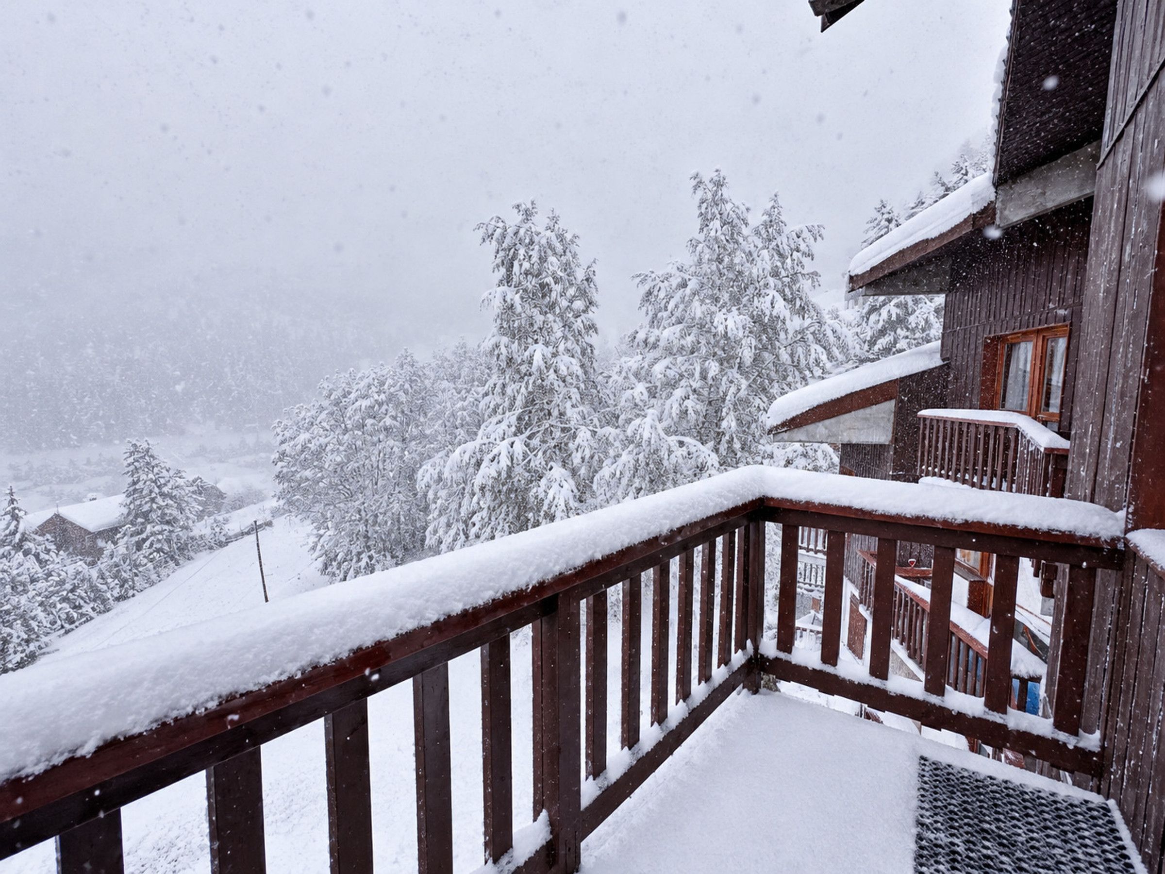 A wooden balcony with snow-covered railings overlooking a snowy landscape with trees and buildings.