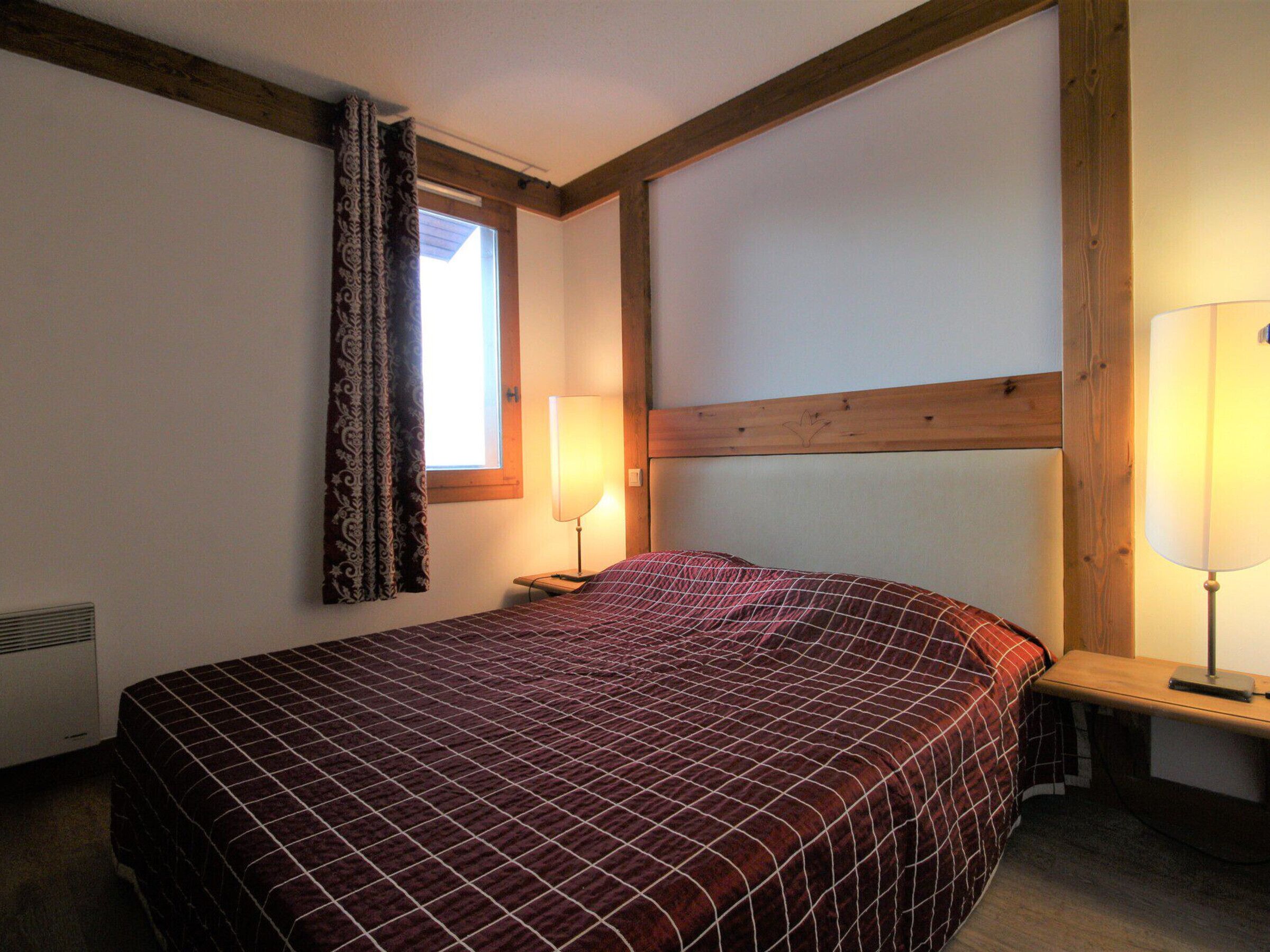 A bedroom with a bed, two bedside lamps, a window with curtains, and wooden beams on the ceiling.
