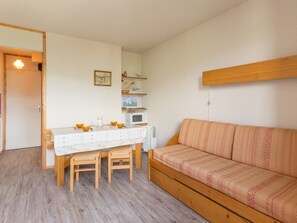 Un espacio habitable pequeño con una mesa de comedor de madera, dos sillas de madera, un sofá, un microondas y una pequeña estantería con objetos decorativos.