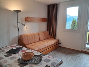 Une pièce comprenant un canapé, une table avec un carnet et un bol, une lampe, et une fenêtre donnant sur un paysage montagneux.