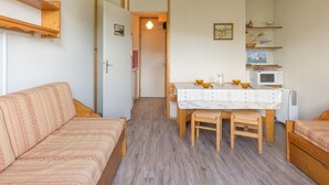 Ein kleiner, gemütlicher Raum mit Holzboden, einem Bett mit gemustertem Überwurf, einem Holztisch mit weißem Tischläufer und einer Wandküche.