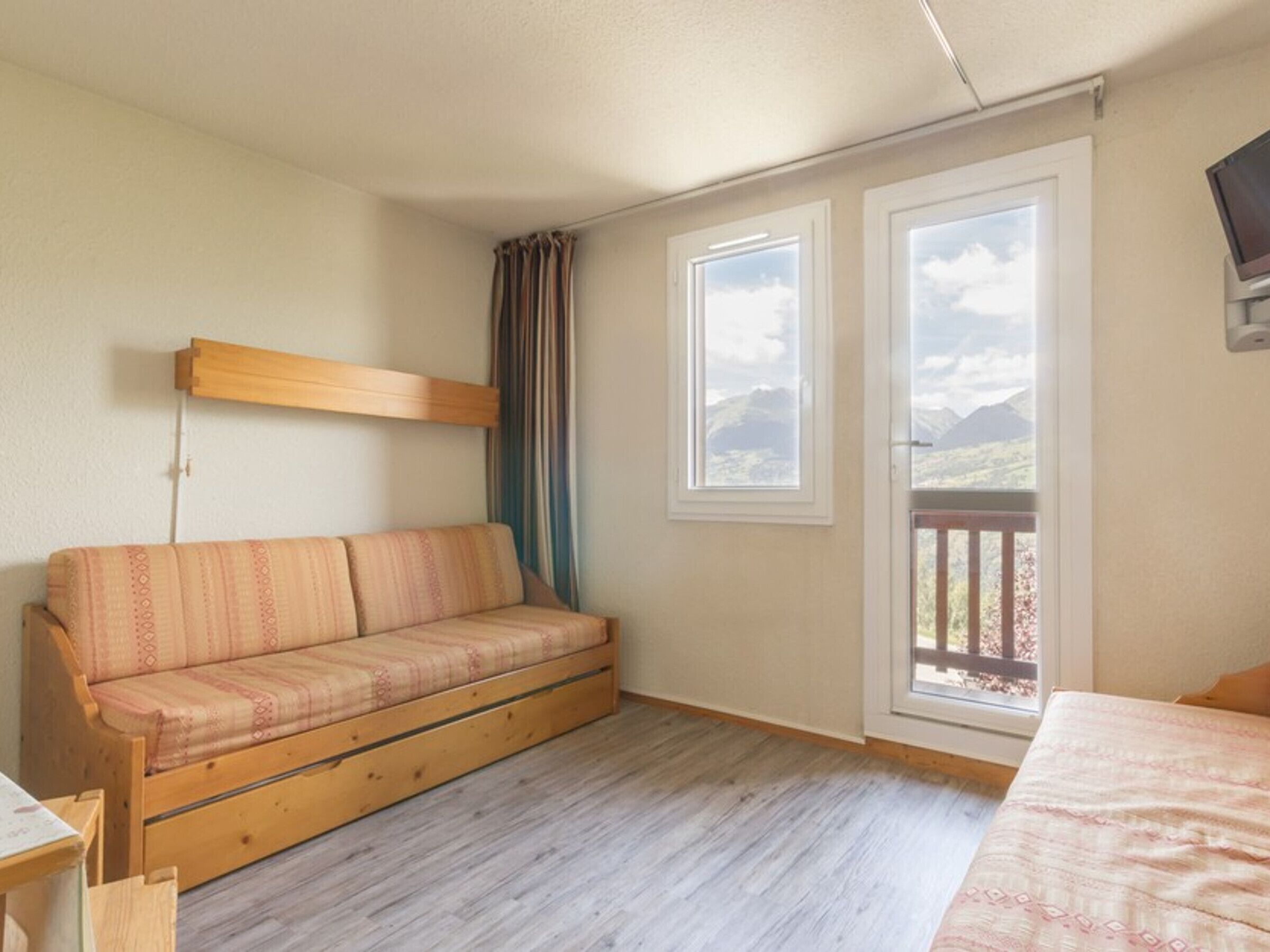 A room with a wooden sofa, two windows with mountain views, a TV mounted on the wall, and a wooden floor.