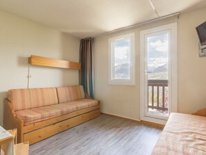 Una habitación con sofá de madera, dos ventanas con vistas a la montaña, un televisor montado en la pared y suelo de madera.