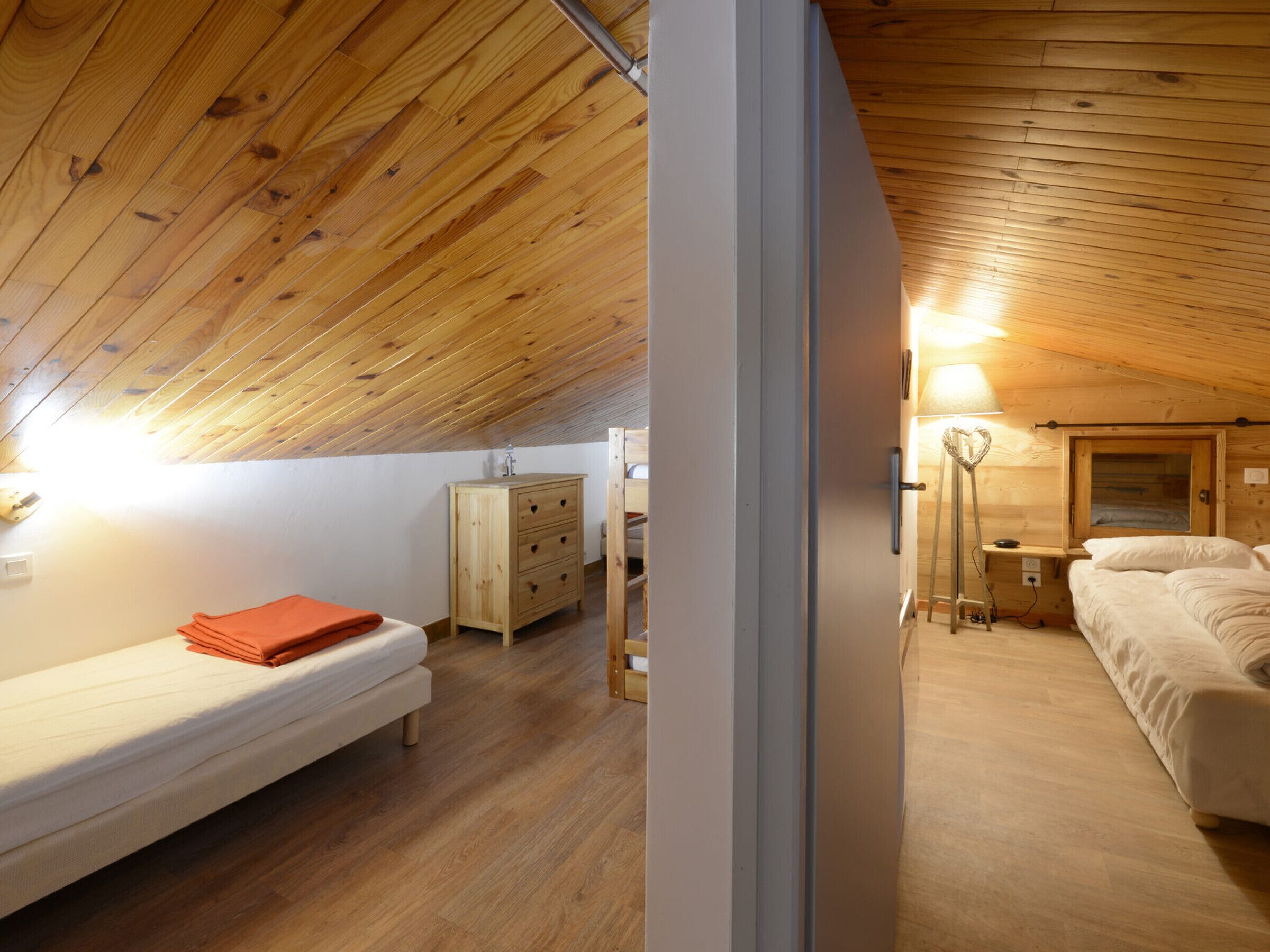 Ein gemütliches Schlafzimmer im Dachgeschoss mit Holzbalkendecke, einem Bett, einem Nachttisch, einer Kommode und einem Schreibtisch mit Lampe.