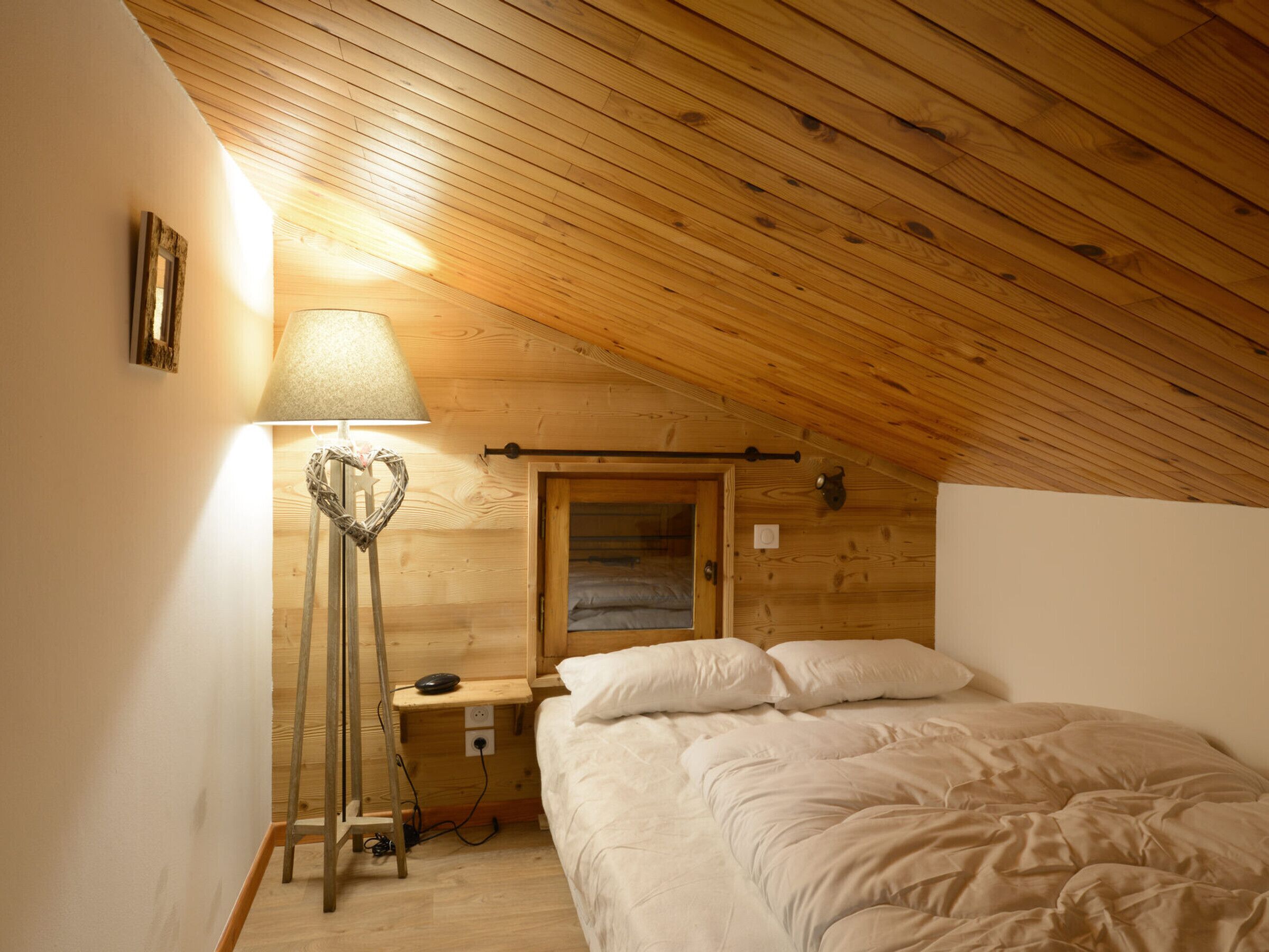 A cozy attic bedroom with a sloped wooden ceiling, a bedside lamp, a small window, and a neatly made bed.
