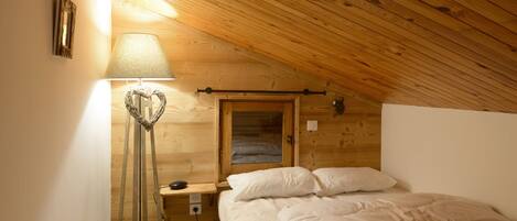 A cozy attic bedroom with a sloped wooden ceiling, a bedside lamp, a small window, and a neatly made bed.