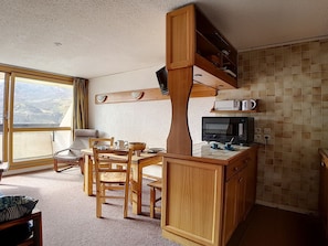 A compact kitchen with wooden cabinets, a dining table with chairs, a microwave, and a large window offering a view. - Cozy duplex at the foot of the slopes with balcony (Les Menuires)